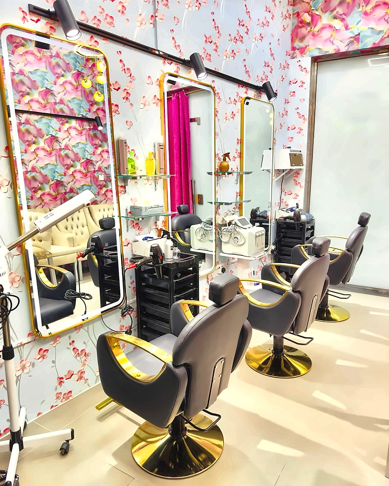 Row of black-and-gold styling chairs facing gold-framed lit mirrors against the salon's pink, turquoise and blossom wallpaper