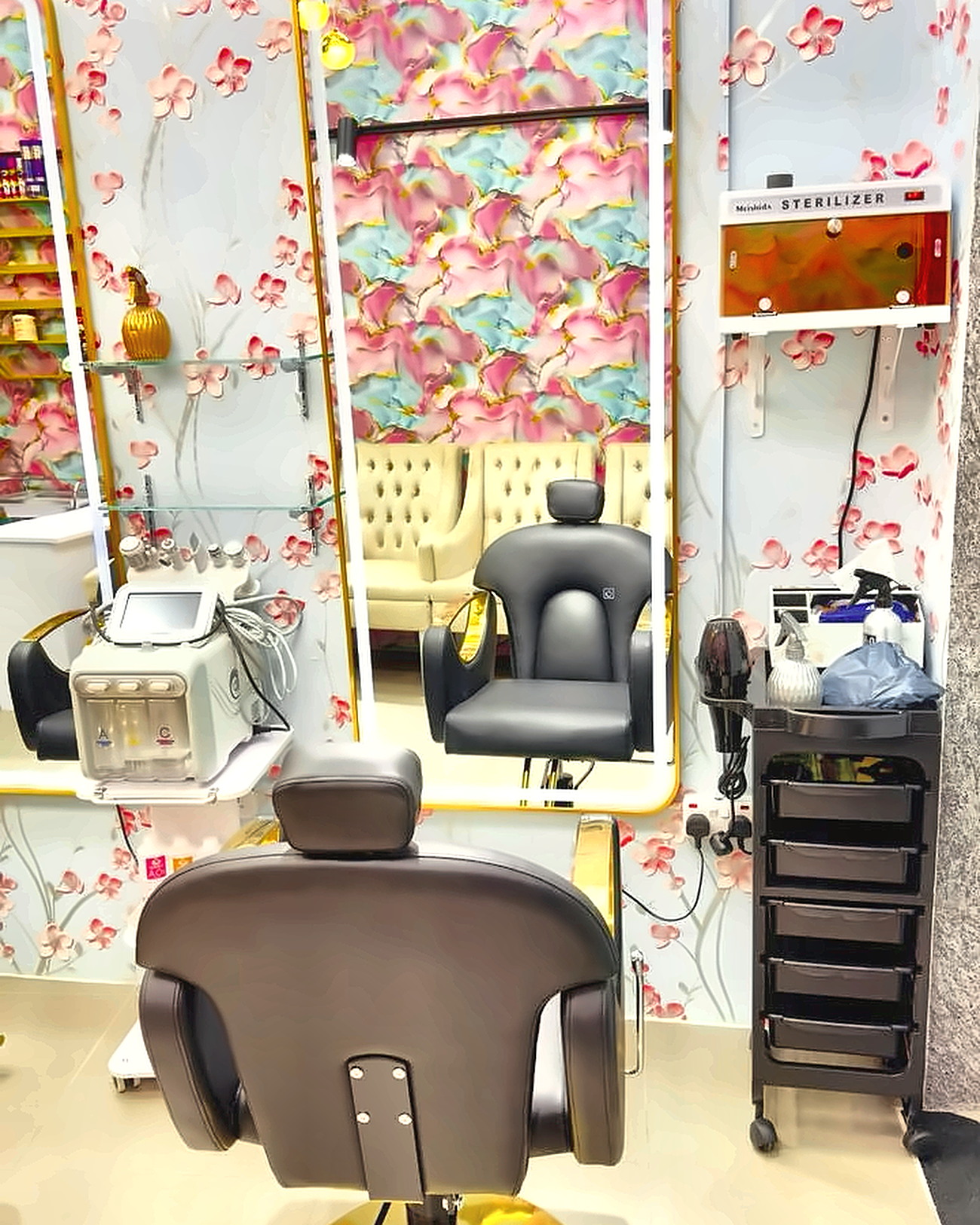 Styling chair facing a gold-framed lit mirror, with the wall-mounted tool sterilizer and trolley beside it