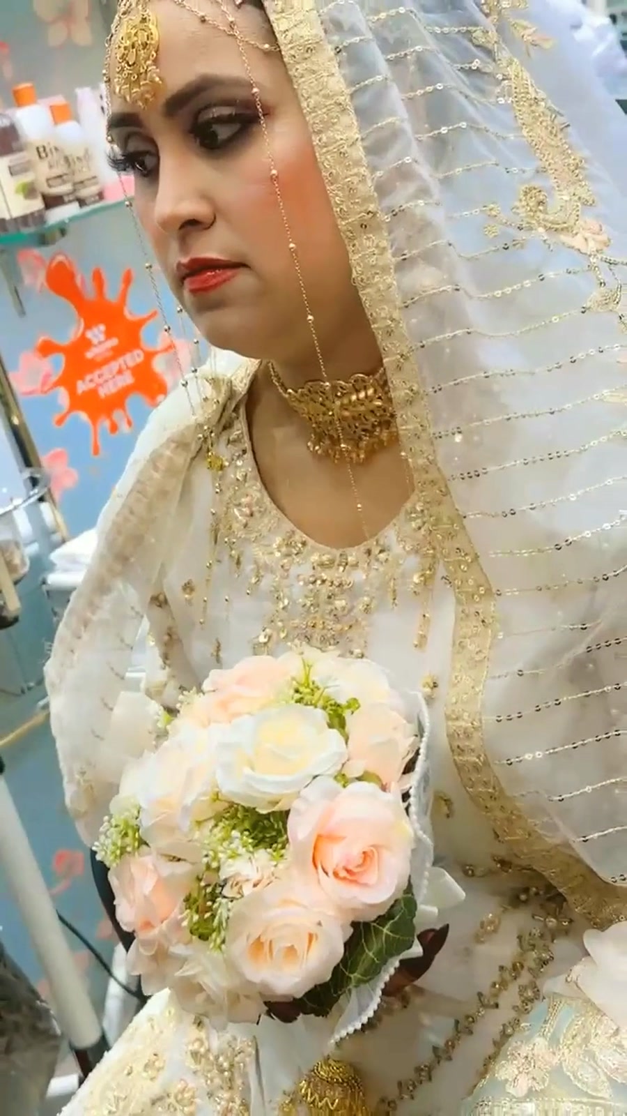 Bride in an ivory and gold gown with a gold choker and maang tikka, holding a cream rose bouquet in the salon